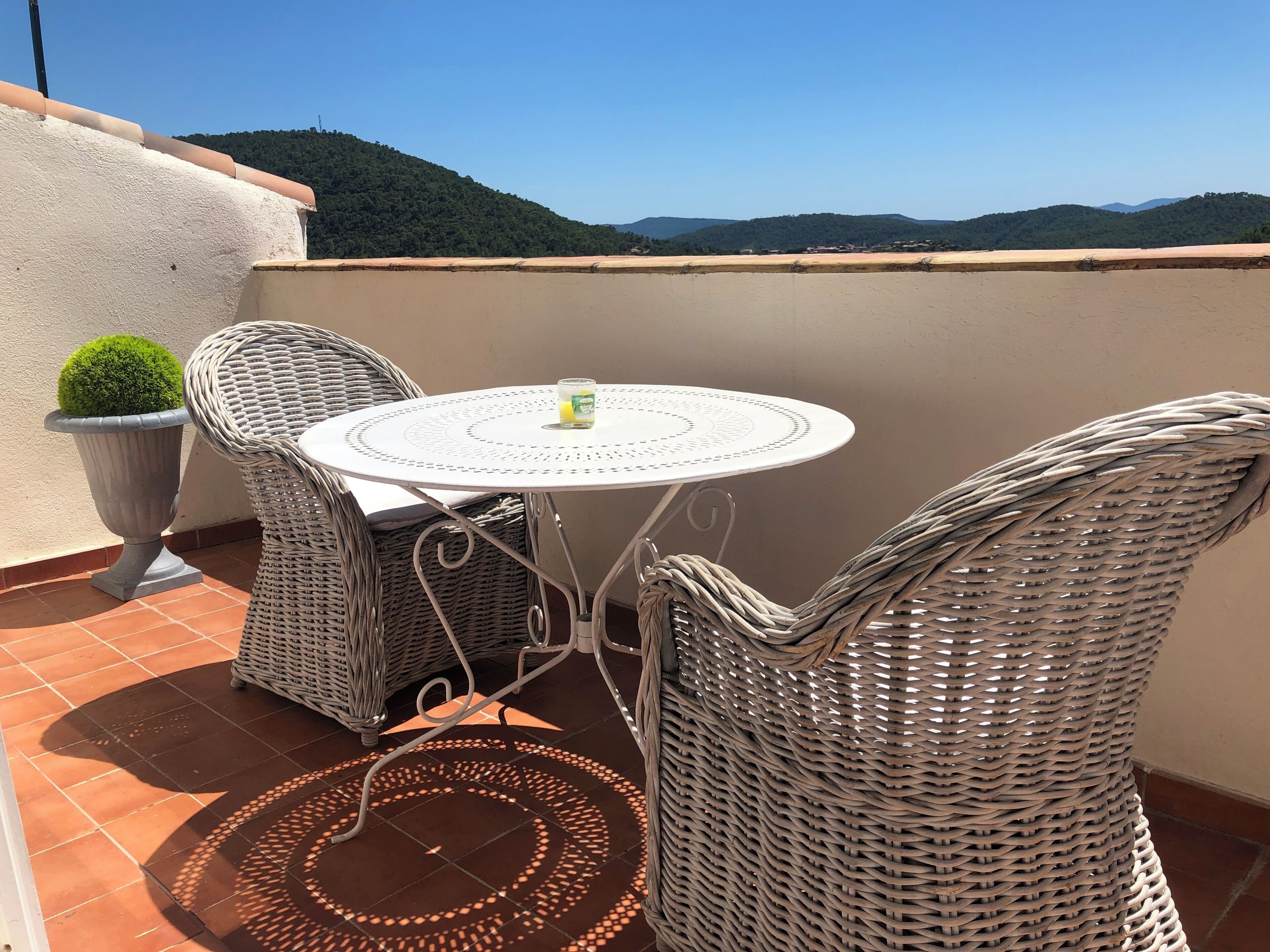 Un petit espace extérieur aménagé avec une table ronde et des chaises en osier, un pot de fleurs et une vue sur des collines lointaines.