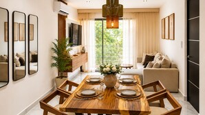 Une salle à manger moderne avec une table en bois dressée pour quatre personnes, entourée de chaises, d’un grand miroir et d’un téléviseur.