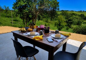 Mesa de jantar ao ar livre com comida e bebidas, rodeada por cadeiras, com uma paisagem campestre como pano de fundo.