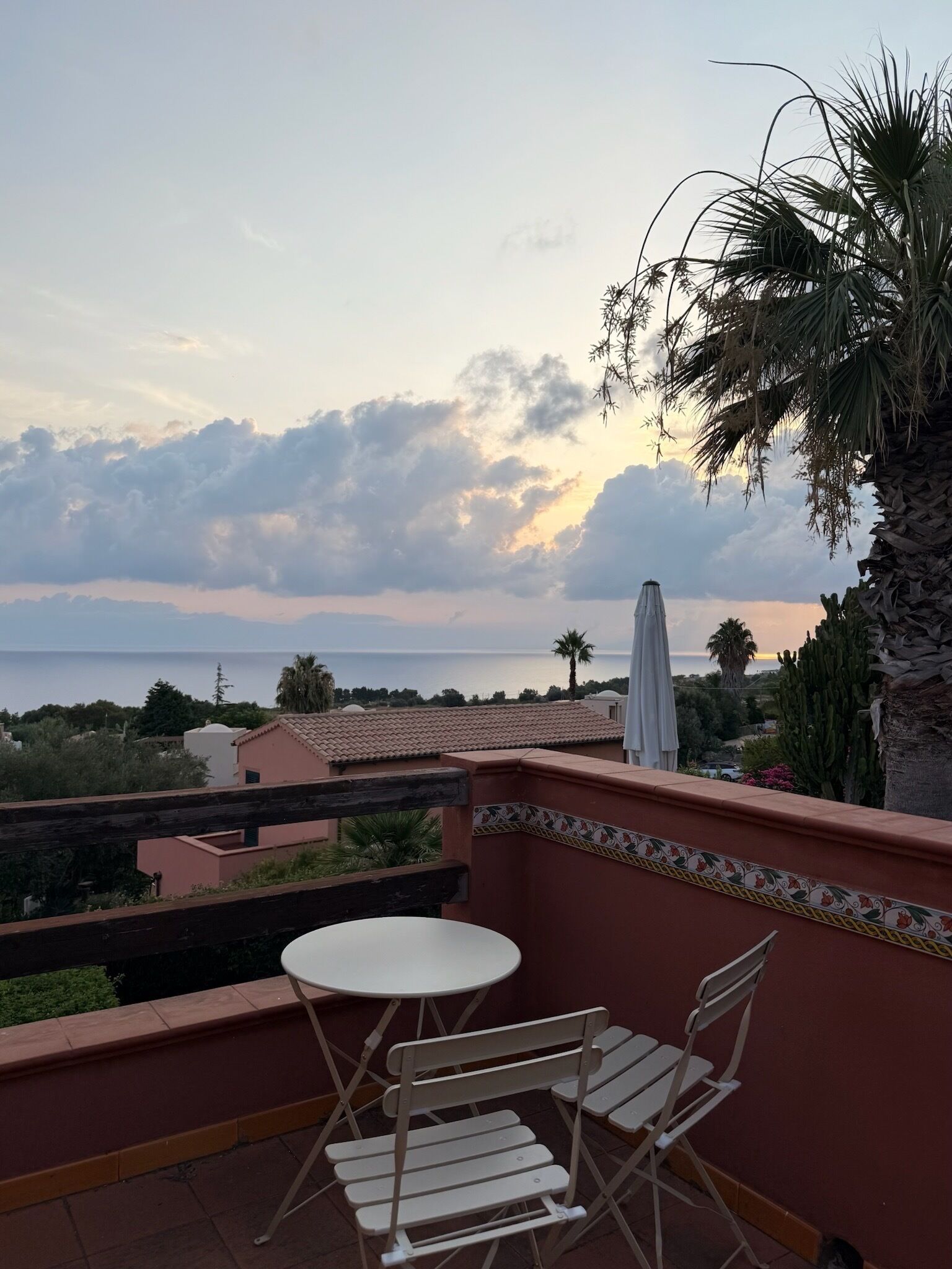 A balcony with a table and chairs, overlooking a coastal view with palm trees and a clear sky.