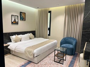 A modern hotel room with a large bed, a blue armchair, a small table, and a nightstand.