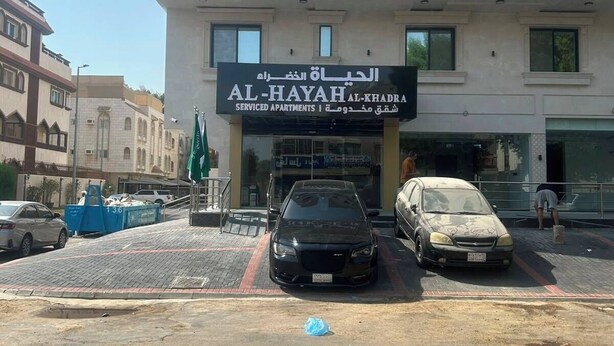 A multi-story building with a sign for 'AL-HAYAH AL-KHADRA' indicating serviced apartments. There are cars parked in front, and a blue trash bin is visible on the sidewalk.