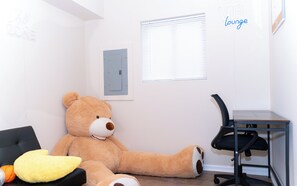 Ein Zimmer mit einem großen Teddybären, einem gelben Kissen, einem schwarzen Bürostuhl und einem Fenster mit Rollläden.