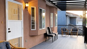 Un espacio al aire libre cubierto con una terraza de madera, dos sillas de mimbre, una puerta blanca con ventanas de celosía y una lámpara fijada a la pared.