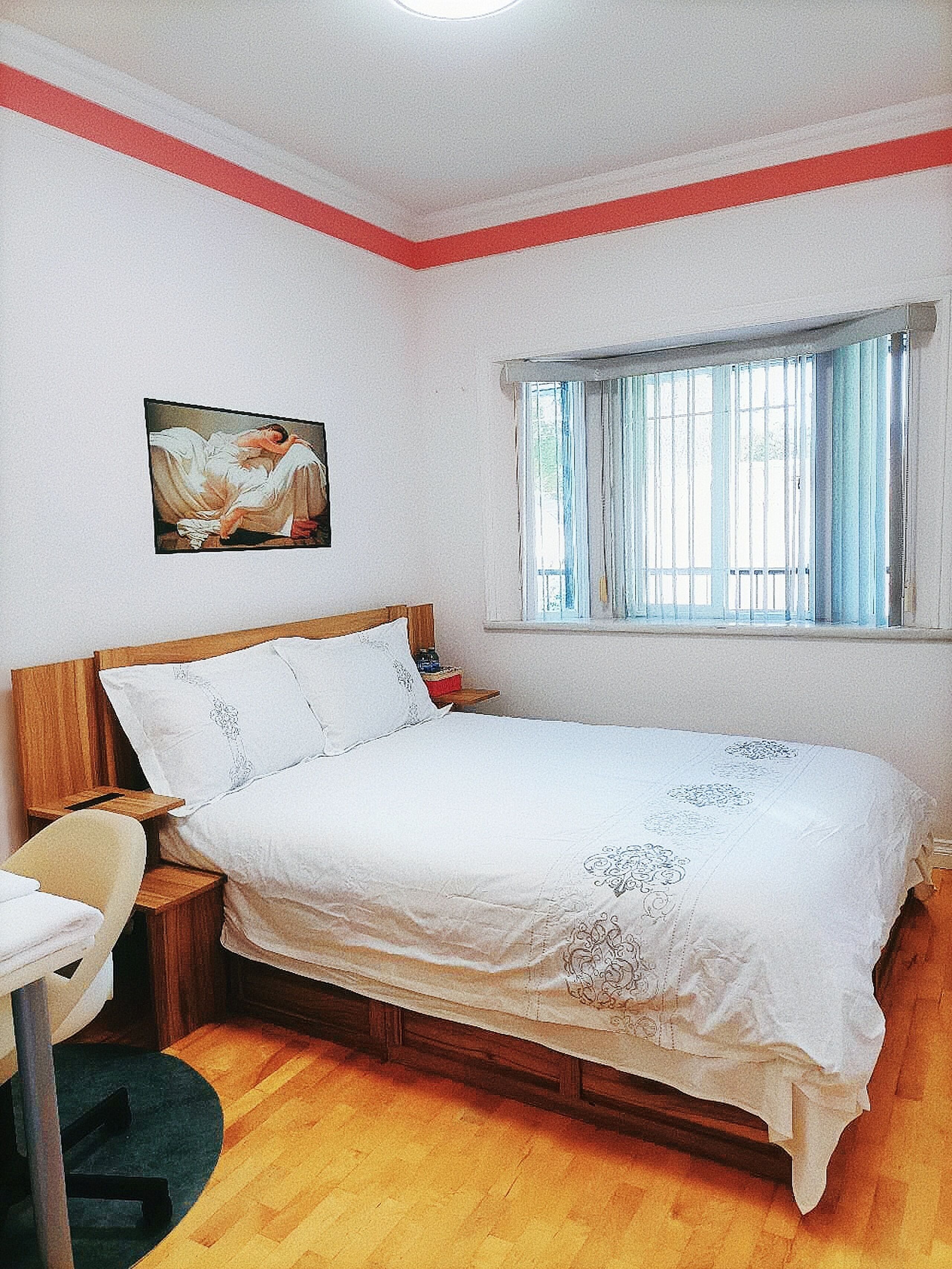 Een slaapkamer met een houten bed, nachtkastje, stoel, raam met gordijnen en een schilderij aan de muur.