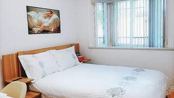 Een slaapkamer met een houten bed, nachtkastje, stoel, raam met gordijnen en een schilderij aan de muur.