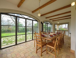 A dining area with a long table, multiple chairs, and large windows offering a view of a garden. - Villa delle Cese (Campagnano di Roma)