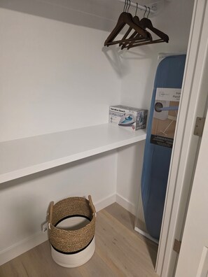 Ein Zimmer mit weißer Wand, einem Kleiderhaken aus Holz, einem Korb, einem blauen Bügelbrett und einer Box mit der Aufschrift 'Hamilton Beach Steam Iron'.