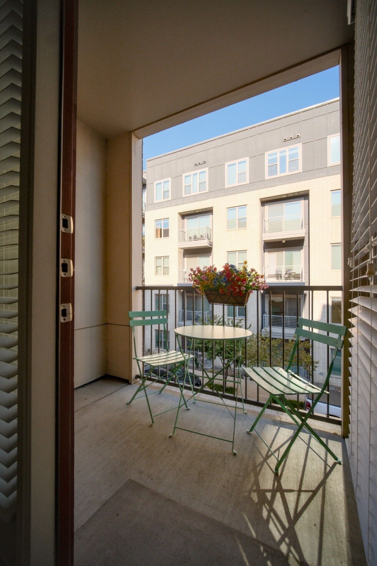 A balcony with a table and chairs, a hanging flower basket, and a view of a multi-story building.
