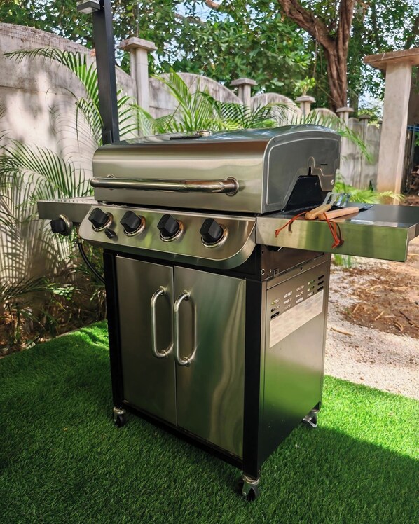 A stainless steel outdoor grill with four burners, a side burner, and a built-in oven, set on artificial grass.