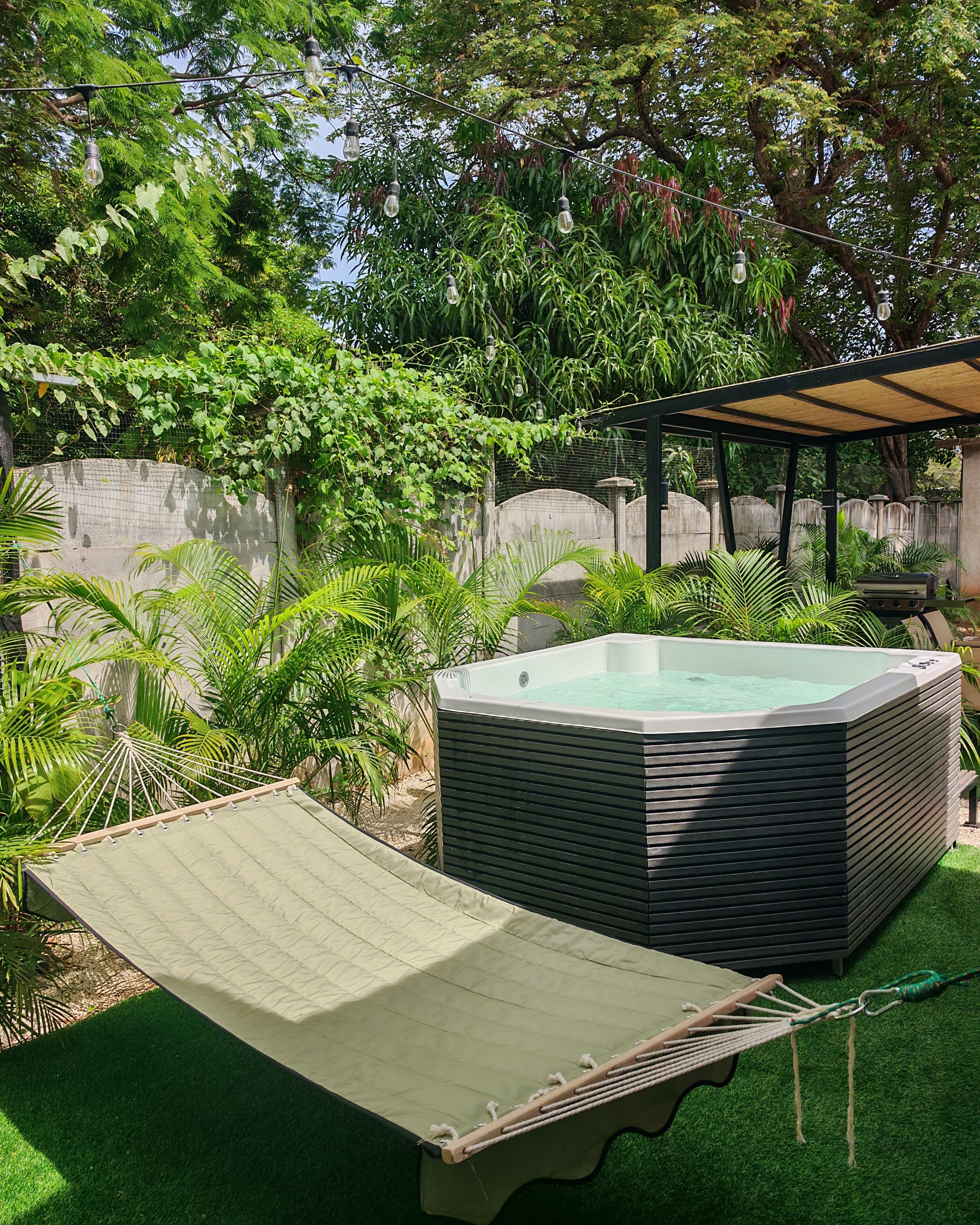 A hot tub with a wooden deck and a hammock in a lush garden.