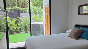 A bedroom with a bed, a sliding glass door to a garden, and a small air conditioning unit.