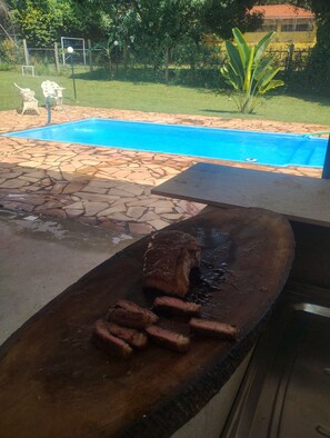 An outdoor barbecue area with a stone-tiled patio, a pool in the background, and a grassy lawn. - Accommodations with a pool table in Igarapé (Igarapé)