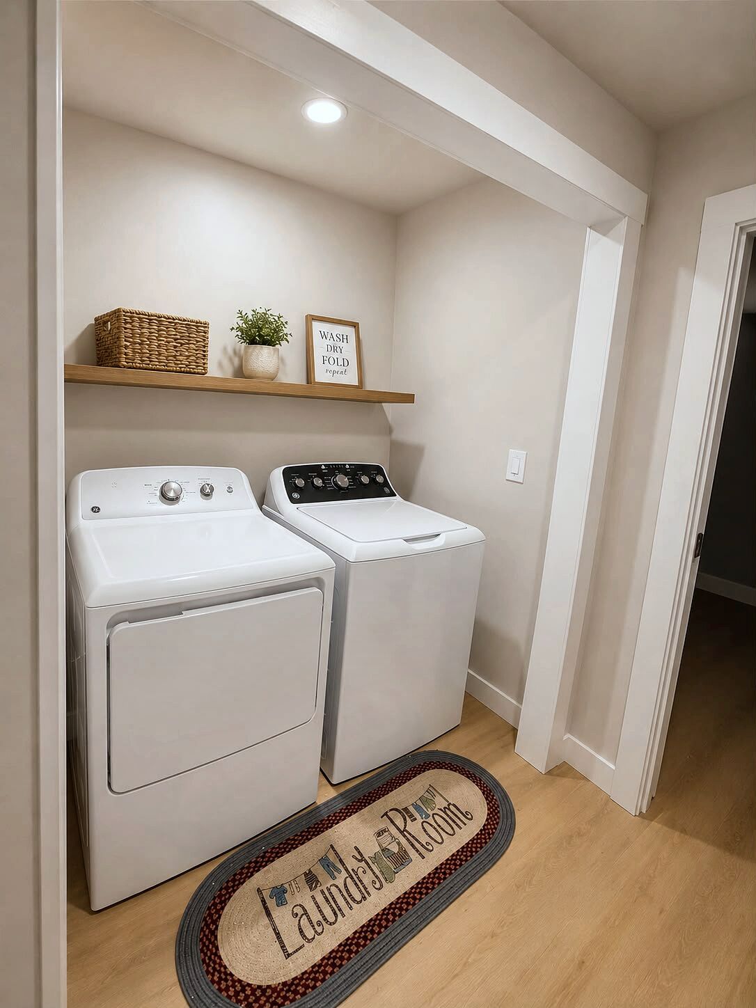 A laundry room with two front-loading washers and dryers, a wooden shelf with a basket and a potted plant, and a welcome mat with 'Laundry Room' text.