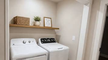 A laundry room with two front-loading washers and dryers, a wooden shelf with a basket and a potted plant, and a welcome mat with 'Laundry Room' text.