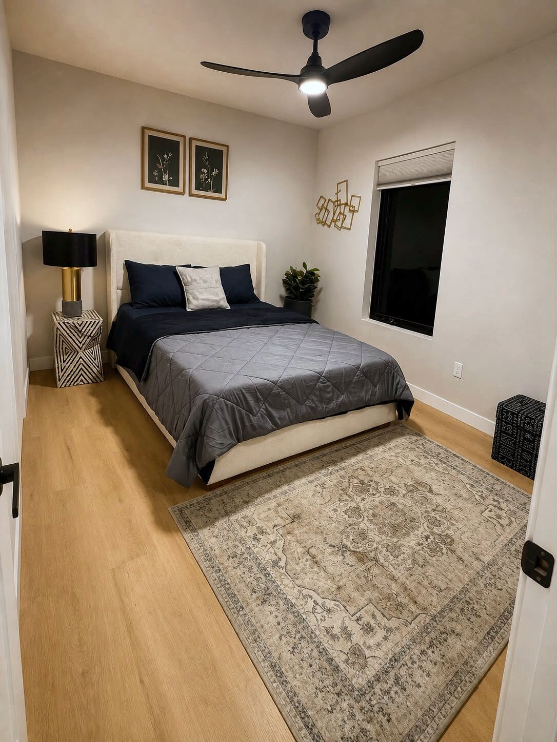 A bedroom with a bed, a ceiling fan, a nightstand, a potted plant, a window, and a patterned rug.