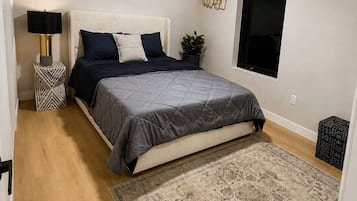 A bedroom with a bed, a ceiling fan, a nightstand, a potted plant, a window, and a patterned rug.