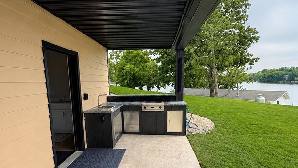 En overdækket terrasse med grill og opbevaringsskabe, en dør ind til en bygning, samt et græsareal med træer og udsigt til søen.