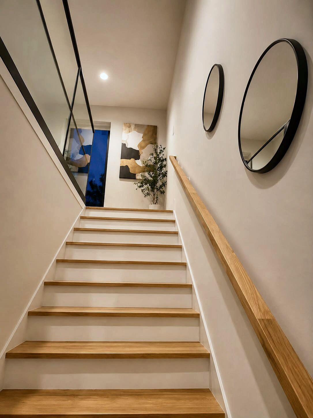 Una escalera con escalones y baranda de madera que conduce a un rellano con un espejo fijado a la pared y un cuadro abstracto enmarcado.