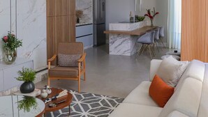 Una sala moderna con un sofá, un sillón y una mesita de centro. Cuenta con una zona de cocina, un refrigerador y una mesa de comedor con sillas.