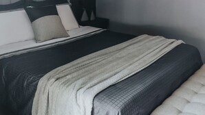 Una cama con una colcha gris acolchada, una manta blanca y un almohadón de color gris.