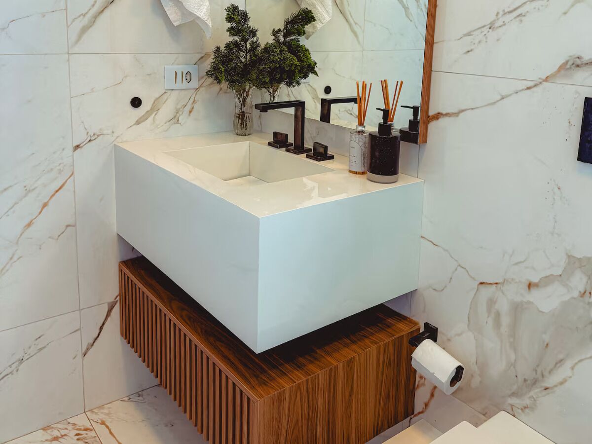 Ein Badezimmer mit einem weißen Marmorwaschbecken, einem Holzlagerraum, einem schwarzen Wasserhahn und einem Spiegel.