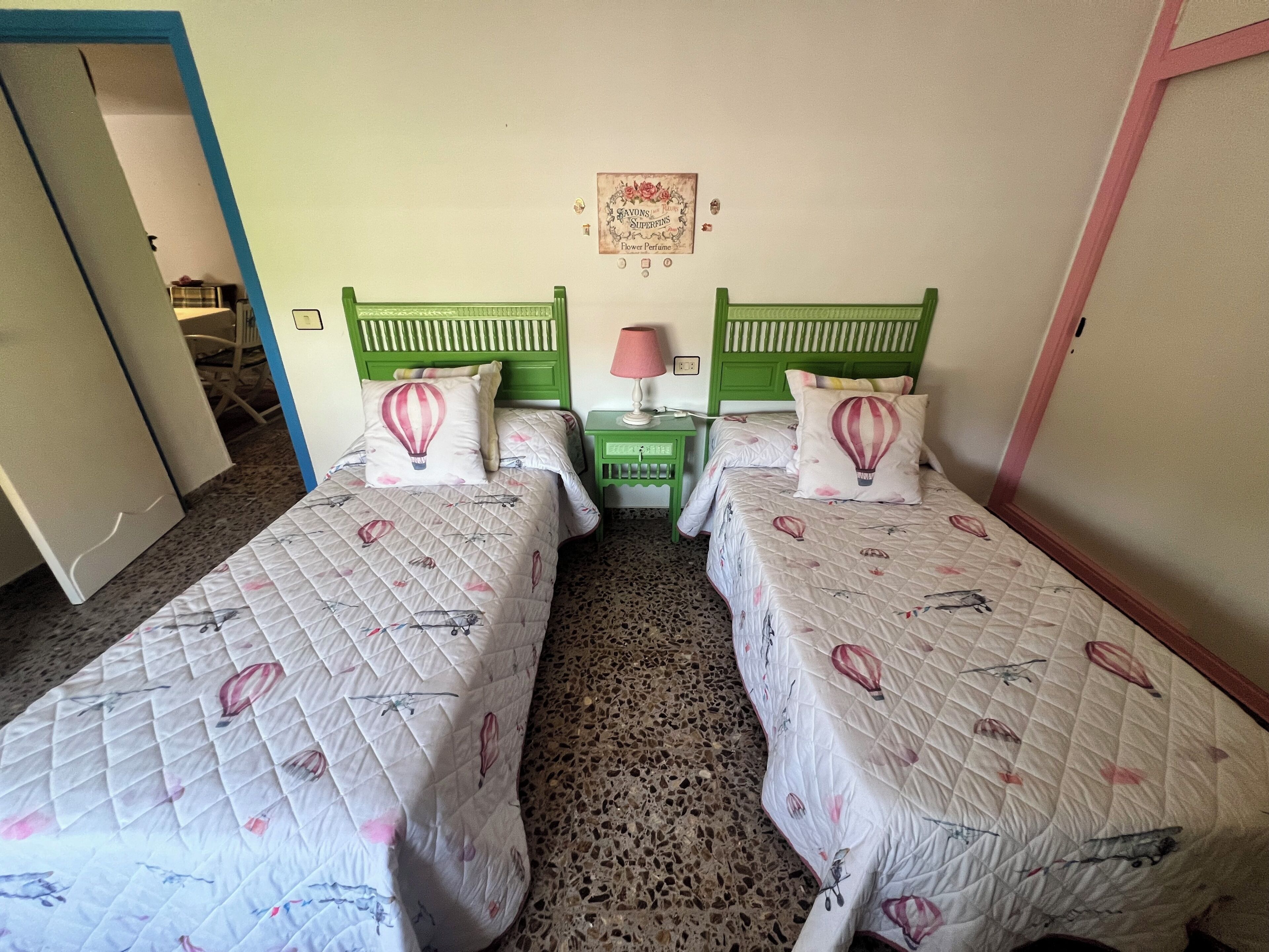 A room with two beds, each with a quilted bedspread featuring a floral pattern. There is a small bedside table with a lamp between the beds.