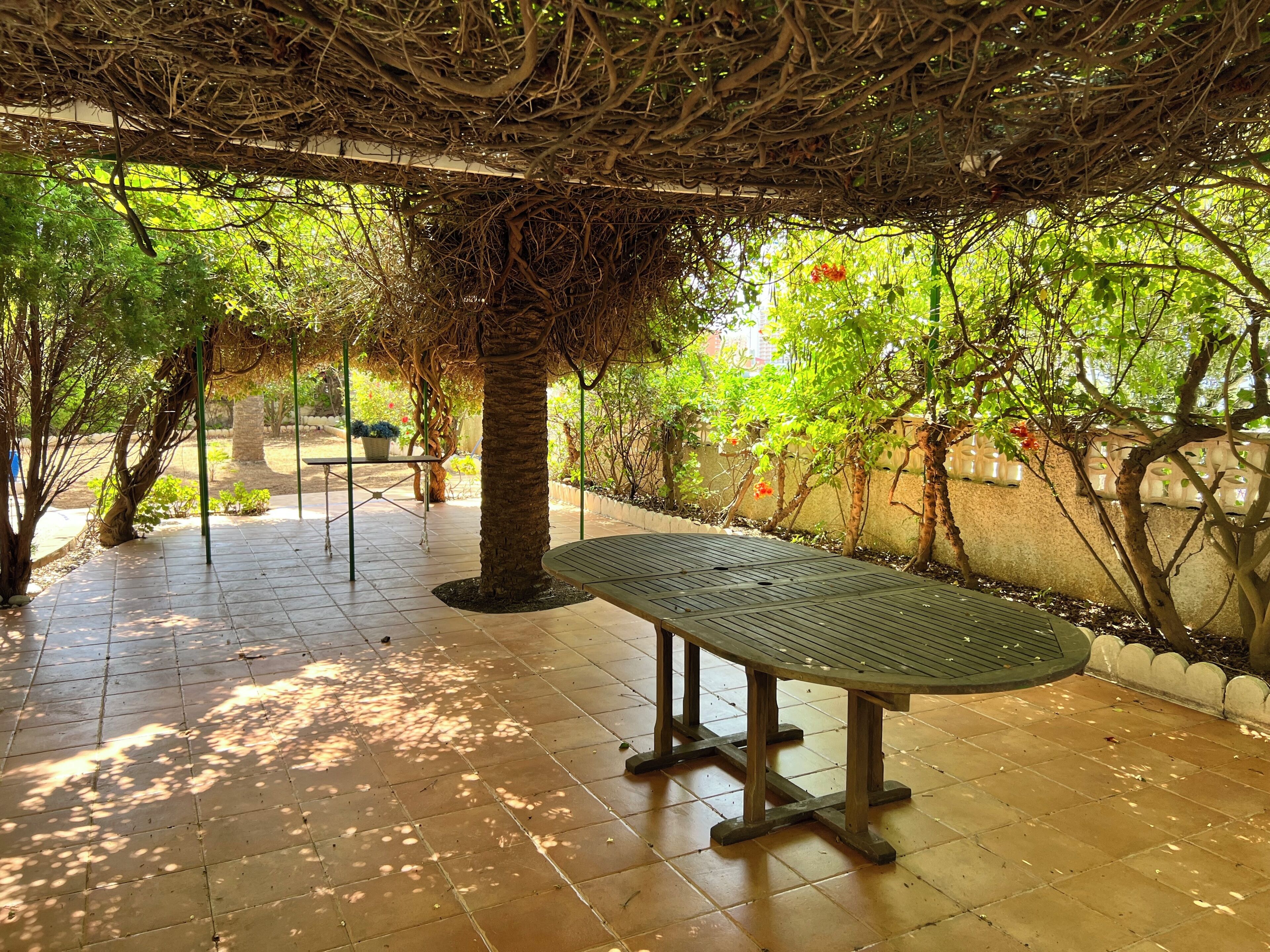 A covered outdoor area with a bench, tiled flooring, and greenery.