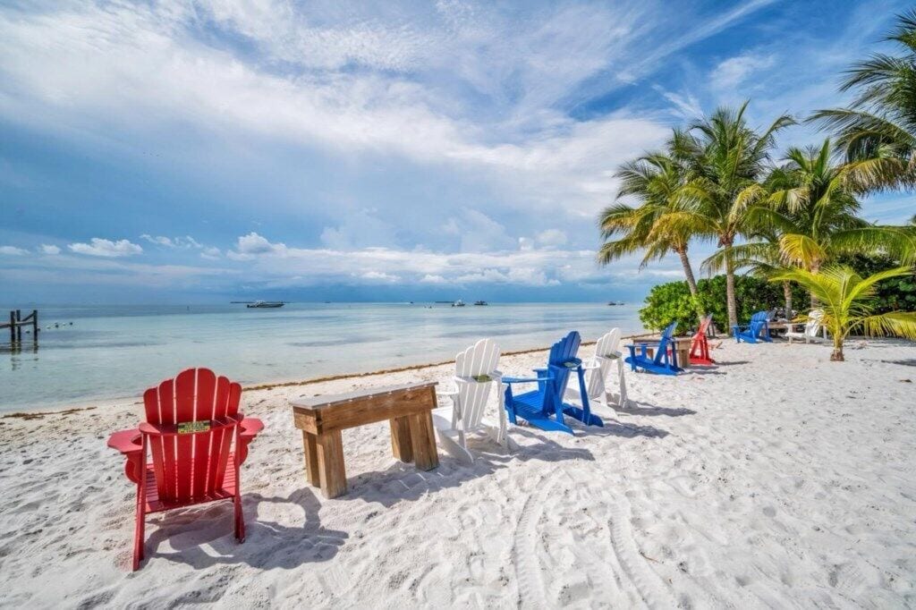 A beach with white sand, a wooden bench, and red and blue Adirondack chairs.
