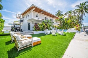 Ein modernes, zweistöckiges Haus mit einer überdachten Terrasse, Sitzgelegenheiten im Freien und üppiger Bepflanzung.