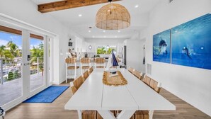 Uma ampla área de jantar com uma mesa branca e cadeiras de madeira, uma grande janela com vista para palmeiras e um teto com vigas aparentes.