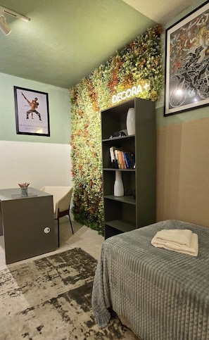 Ein Schlafzimmer mit einem Bett, einem Schreibtisch, einem Bücherregal, einer Wandpflanze und einem gerahmten Bild.