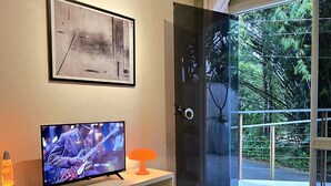 A modern living room with a flat-screen TV, a framed abstract painting, and a glass door leading to a balcony with a view of greenery. - A cozy studio apartment with the charm of an open-concept design that feels like home. (Joinville)