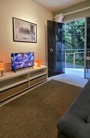 Ein modernes Wohnzimmer mit einem Flachbildfernseher, einem gerahmten abstrakten Gemälde und einer Glastür, die auf einen Balkon mit Blick auf Grünflächen führt.