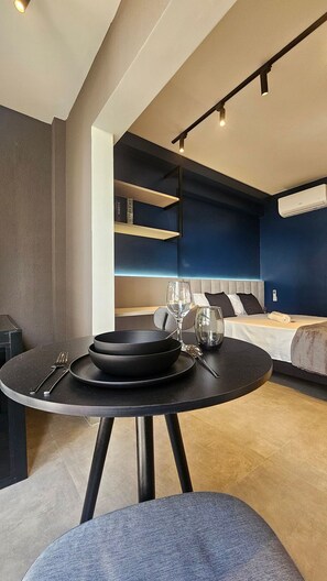 A modern hotel room with a round dining table, a bed with a headboard, and built-in shelving. - Cond. Constantino Campo Belo NR (São Paulo)