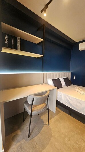 Une petite pièce comprenant un lit, un bureau et une chaise. Des étagères à livres se trouvent au-dessus du bureau.