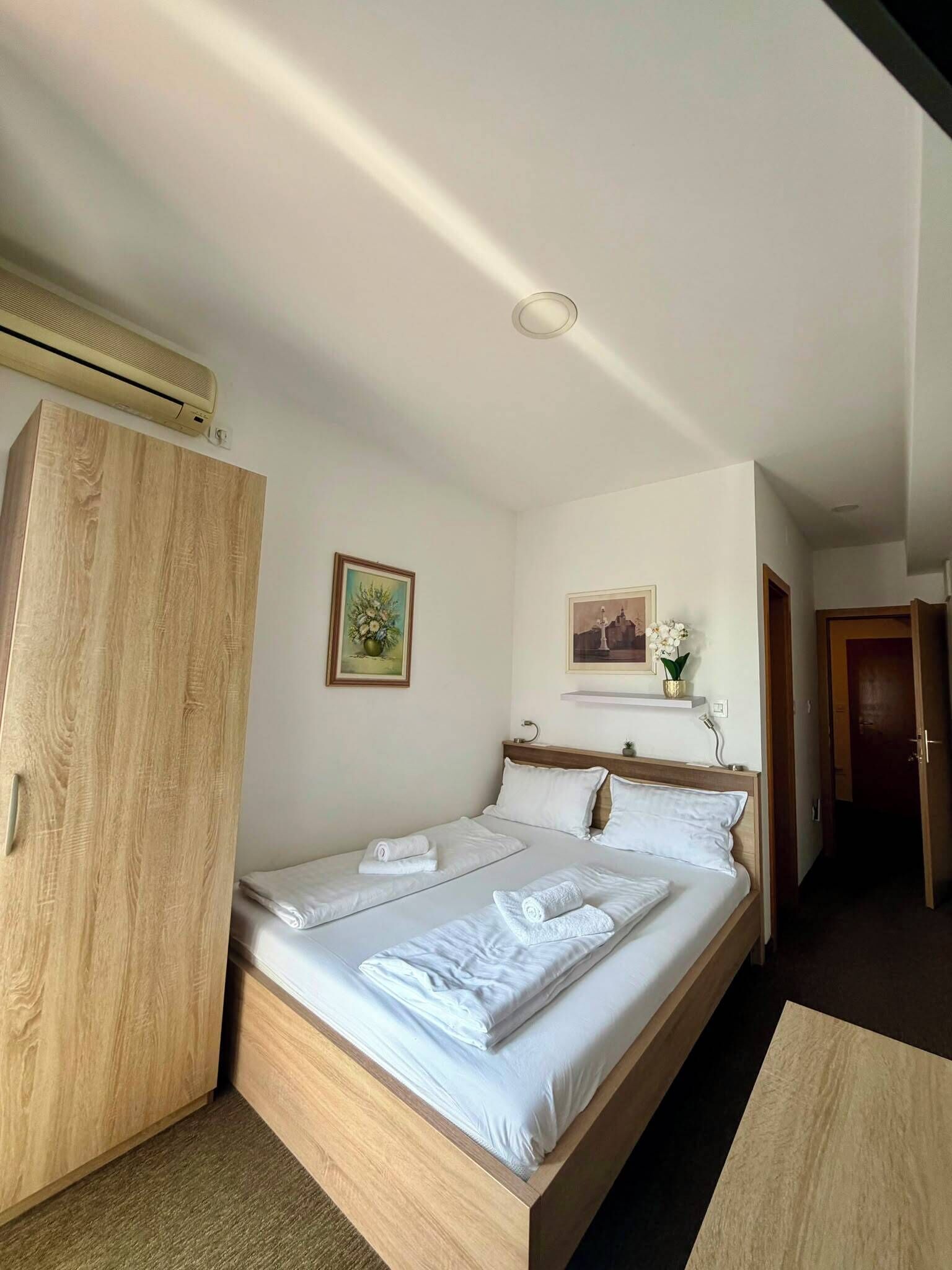 A single bed with white linens, a wooden wardrobe, a small shelf with a vase, and a painting on the wall.