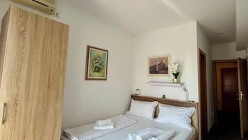 A single bed with white linens, a wooden wardrobe, a small shelf with a vase, and a painting on the wall.