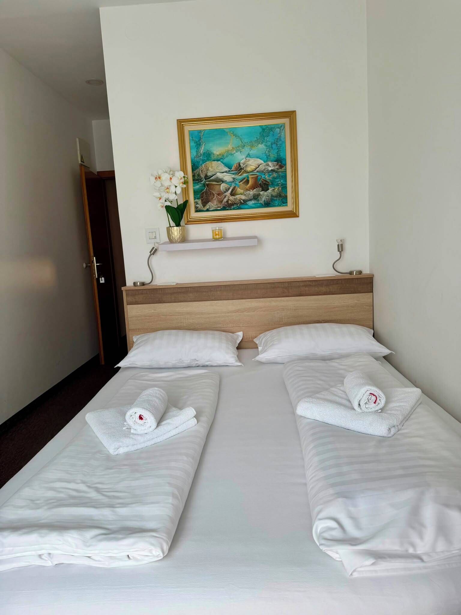 A hotel room with a bed, two pillows, a painting on the wall, a vase with flowers, and a nightstand with a lamp.