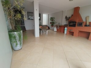 A spacious living area with a tiled floor, a dining table with chairs, a kitchen area with a brick oven, and potted plants.