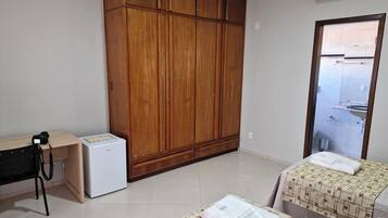 A room with a wooden wardrobe, a small refrigerator, a desk with a chair, and two beds with towels.
