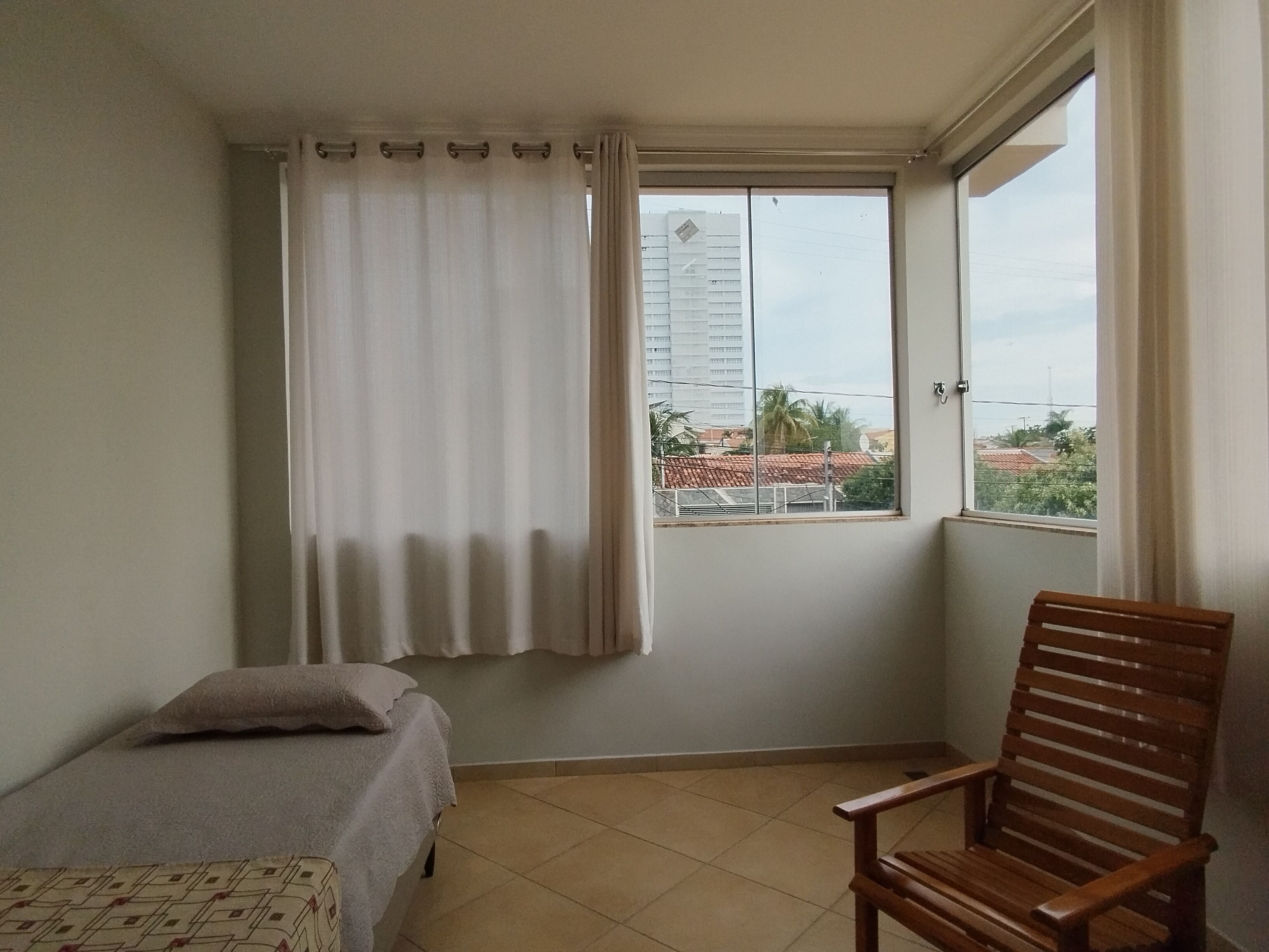 Una habitación con cama, una silla de madera, un ventanal con cortinas y vistas a edificios.