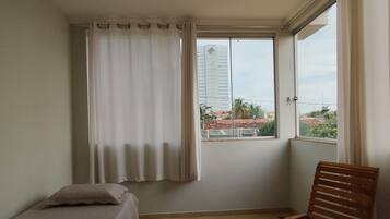 Una habitación con cama, una silla de madera, un ventanal con cortinas y vistas a edificios.