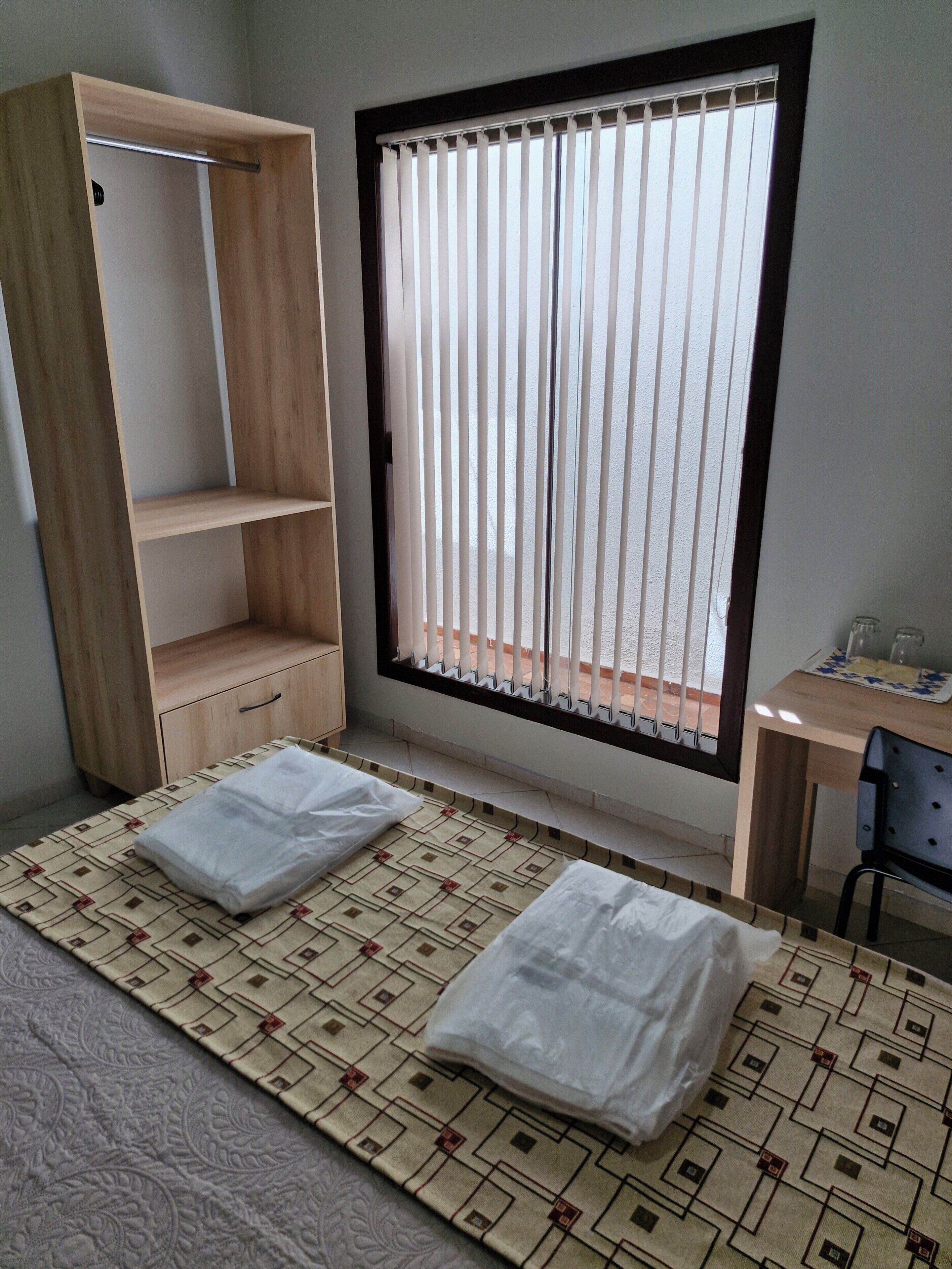 A room with a bed, a wooden wardrobe, a window with blinds, and a desk with a chair.