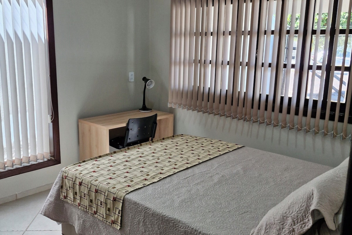 Een slaapkamer met een bed, nachtkastje, bureau en raam met jaloezieën.