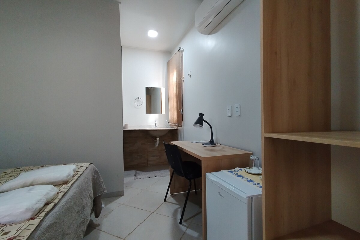 Een kleine kamer met een bed, een bureau, een stoel, een kleine koelkast en een badkamer op de achtergrond.