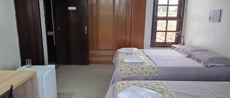 A hotel room with two beds, a wooden wardrobe, a desk with a chair, a small refrigerator, and a window with a view.