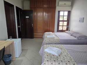 A hotel room with two beds, a wooden wardrobe, a desk with a chair, a small refrigerator, and a window with a view.
