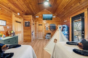 Ein Blockhütten-Innenraum mit zwei Betten, einem Deckenventilator, einem Feuerlöscher, einer Holzwand mit einem Bärenbild, einem kleinen Hausmodell und einem Zelt.