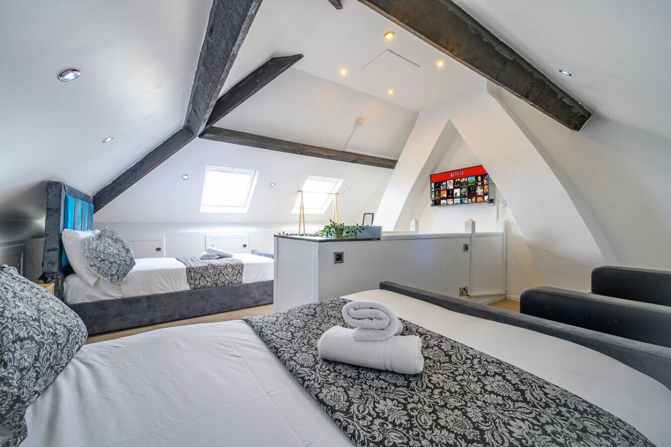 A modern loft bedroom with a sloped ceiling, a bed with patterned bedding, a small kitchenette, and a TV mounted on the wall.